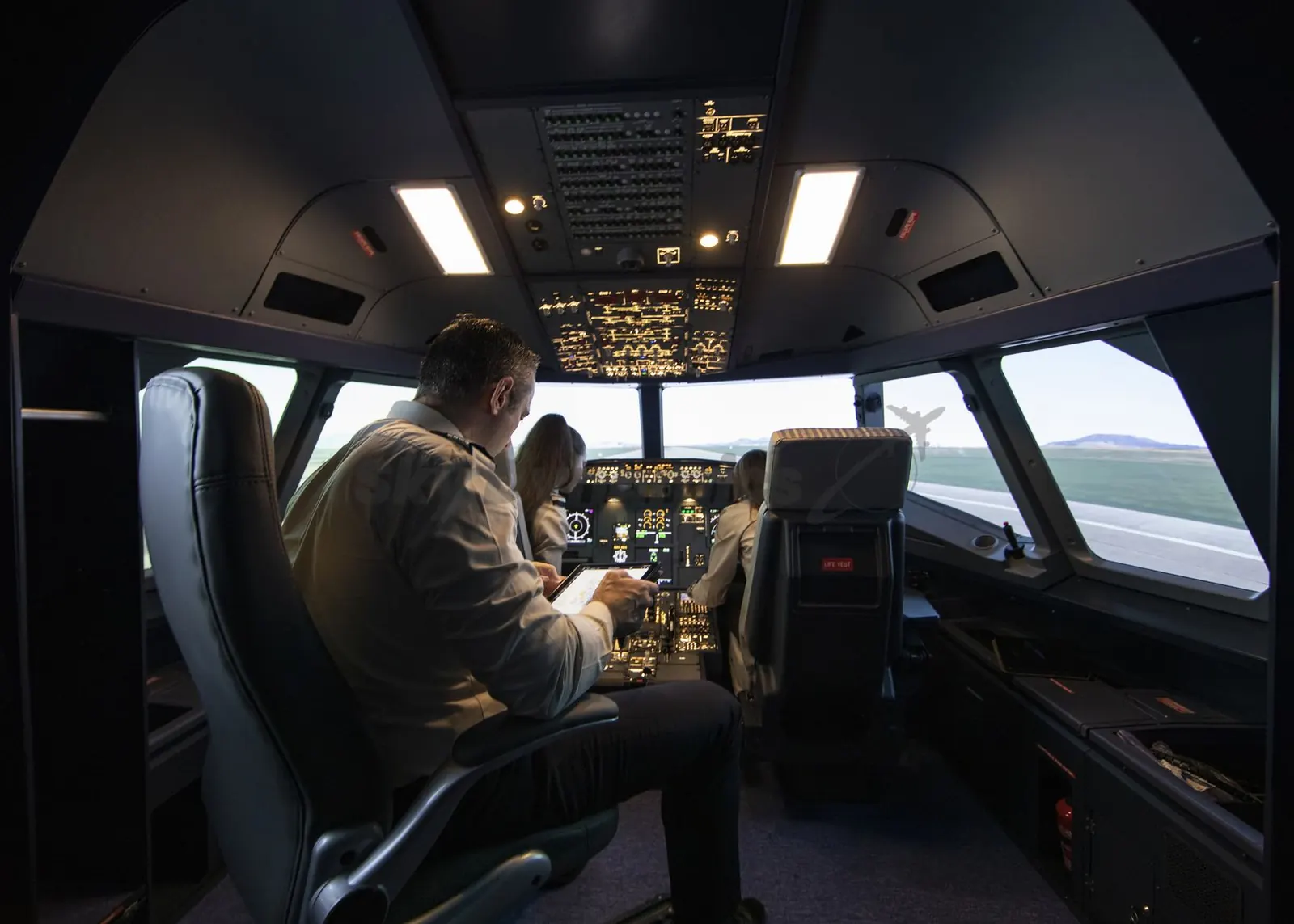 An instructor seated behind two trainees, holding a tablet, during a session in a SkyDynamics A320 FNPT II simulator