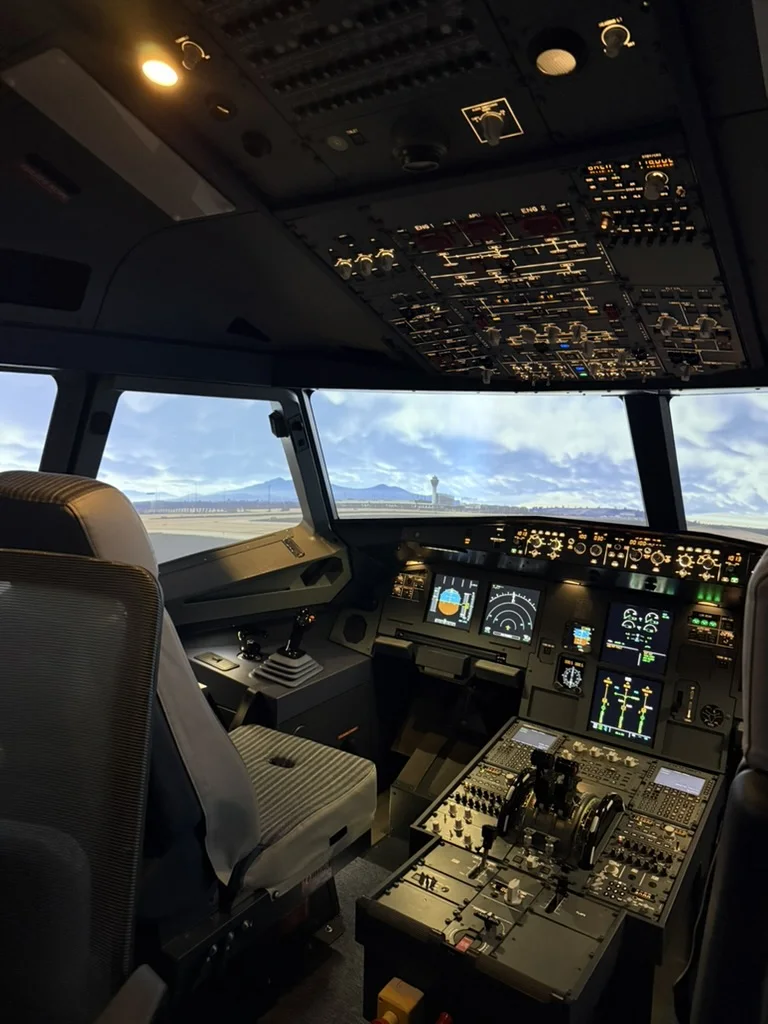 Captain's side of the A320 FNPT II simulator on the ground at an airport, with the overhead panel lit